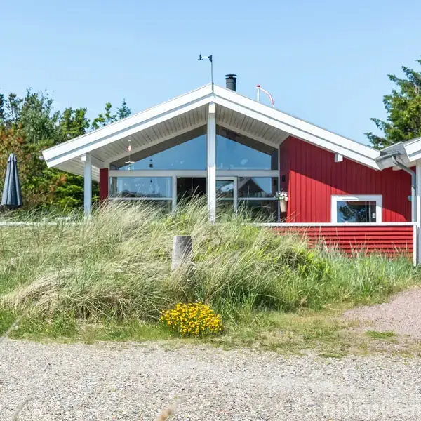 Rødt træhus med store vinduer og hvidt tag, omgivet af græs og vilde blomster, står i en naturpræget have med træer i baggrunden.