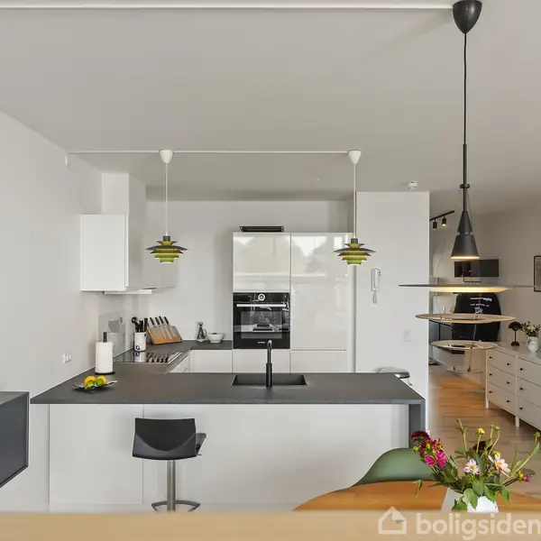 Køkken med moderne ø og sort barstol, lamper hængende over bordet, omgivet af hvide skabe og lyse træmøbler. Blomster står på bordet, og væggen har billedkunst.