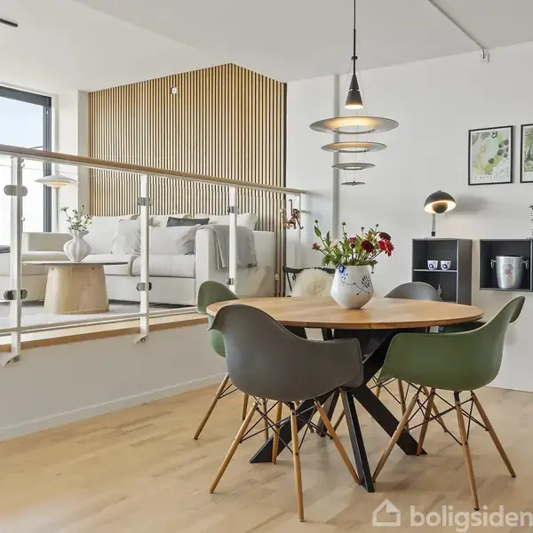 Rundt spisebord med moderne stole står i en lys spisestue. Ovenfor en let forhøjning er en sofasektion med trævægpaneler og store vinduer. Lampen hænger over bordet, og billeder hænger på væggen.