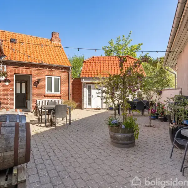 To gårdhuse med rødt tegltag omkranser en brolagt gårdhave, hvor der står et bord og stole til spisning. Krukker med planter og en vintønde skaber en hyggelig atmosfære.