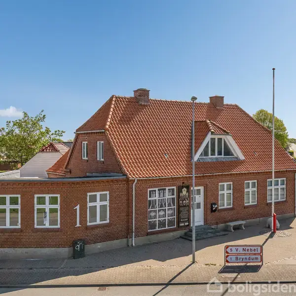 En rød murstensbygning med rødt tegltag står ved en vej. Ved bygningen ses vejskilte med teksten "S.V. Nebel" og "Bryndum". Omkring bygningen er der grønt løv og blå himmel.