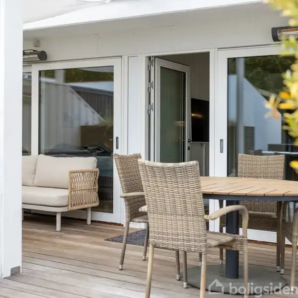 Terrassemøbler står på en overdækket terrasse med trægulv. To stole rundt om et rundt bord nær en sofa med puder, omgivet af planter. Døre åbner ind til huset.