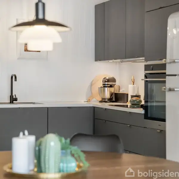 Et moderne køkken med grå skabe, hvide vægge og et hvidt køleskab. En bordplade har en køkkenmaskine, og der hænger en lampe over et rundt spisebord med stearinlys og dekoration.