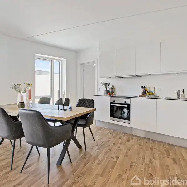 En moderne køkken-spisestue med hvidt køkkenmodul og indbygget ovn. Et træbord omgivet af seks grå stole står til venstre. Rummet er lyst med trægulv og et vindue.