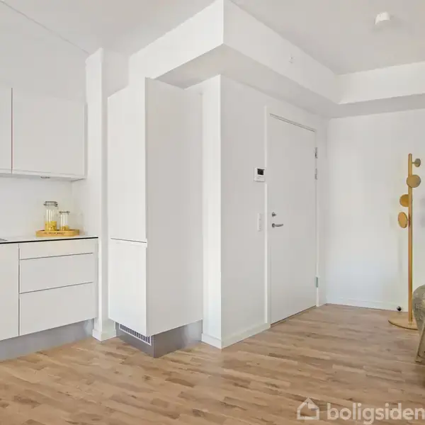 Et lyst køkken med hvide skabe og en bordplade, hvor der står en citrusfrugtskål og glas. Trægulv strækker sig til en entre med dør, bøjlestativ og en stol.