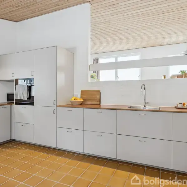 Et moderne køkken med grå skabe indbygget i en L-form. En håndvask står ved et vindue. Brune flisegulve og træloft skaber en enkel og lys atmosfære.