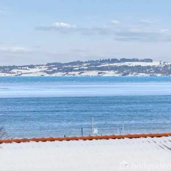 Taget med en skorsten står i forgrunden, mens havet strækker sig mod fjerne, snedækkede bakker under en blå himmel.