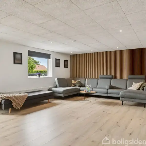 Stue med en stor grå sofa i L-form, placeret mod en væg med træpaneler. Rummet har lyse trægulve, moderne indretning og billeder på væggene. En glasdør fører udenfor.