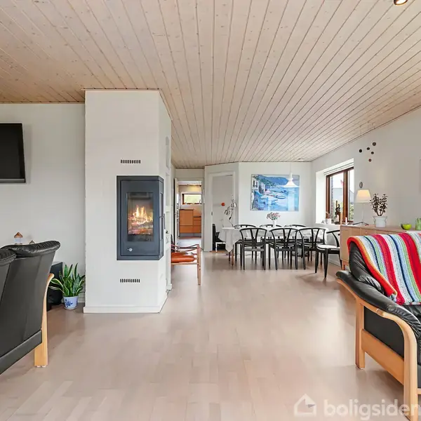 Brændeovn langs en hvid væg opvarmer stue med sorte møbler, spisebord og farverigt tæppe i lyst, træbeklædt rum. Vinduer lader lys strømme ind.