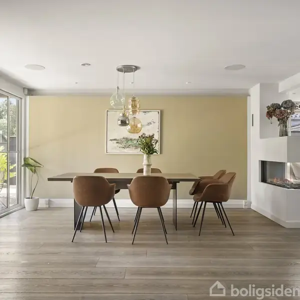 Spisebord omgivet af seks brune stole, placeret i et lyst rum med glasvæg på venstre side og pejs til højre. Der hænger lamper over bordet, og udenfor ses en terrasse.