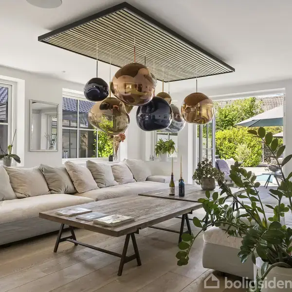 En stor, lys stue med en beige hjørnesofa og et træbord. Dekorative kuglelamper hænger fra loftet. Store vinduer afslører en terrasse og have med grønne planter.