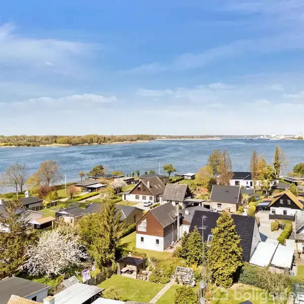 Huse ligger tæt sammen i et boligområde ved en stor blå sø eller fjord, omgivet af træer og grønne haver, under en klar blå himmel.