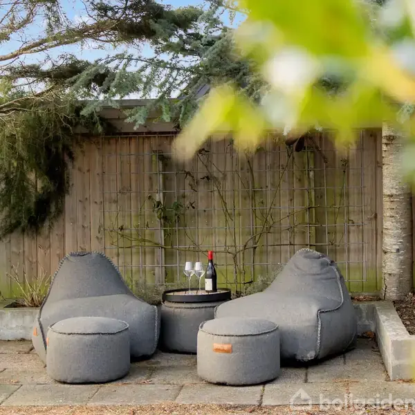 To udendørs loungestole med en flaske vin og to glas på et bord imellem dem, placeret på en terrasse omgivet af træer og et hegn i baggrunden.