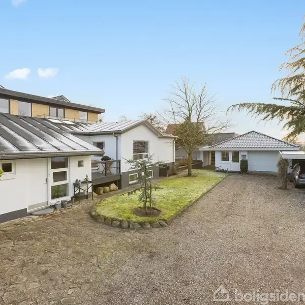 Husene står omgivet af en have med stenbelagt indkørsel. Til højre ses en carport med bil. Himmel og træer danner en rolig baggrund.