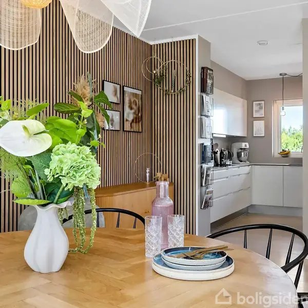 Rundt spisebord med blomster og lamper over sig, omgivet af stole, sofasæt og køkken. Trappe til højre leder til en etage. Lyst og moderne interiør med træ- og metalfinish.
