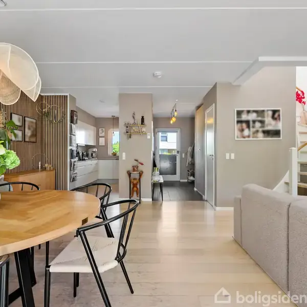 Moderne stue med en sofagruppe til venstre og et spisebord til højre. Store vinduer åbner ud til en grøn have. Væggene er lyse med træpaneldetaljer.