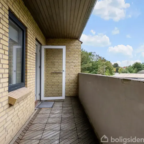 En balkon med træflisegulv og beige murstensvægge. En glasdør står på klem mod facaden. Omkringliggende huse og træer kan ses under en blå skyet himmel.