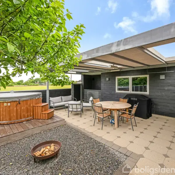 Udendørs terrasse med jacuzzi, brændekurv og spisebord med stole under halvtag. Omkring står et træ og en sofa. Belægning er i fliser og området er indrammet af grønne marker.