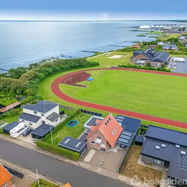 Huse ligger ved en kyst med atletikbane og havudsigt i baggrunden. De omkringliggende grønne områder og boliger danner en fredelig, kystnær byzone.