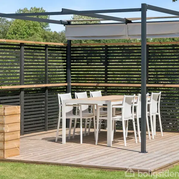 En udendørs terrasse med et bord og seks hvide stole står på et trædæk. Omringet af mørke trælægter, er der blomsterkummer på jorden. Grønne træer ses i baggrunden.