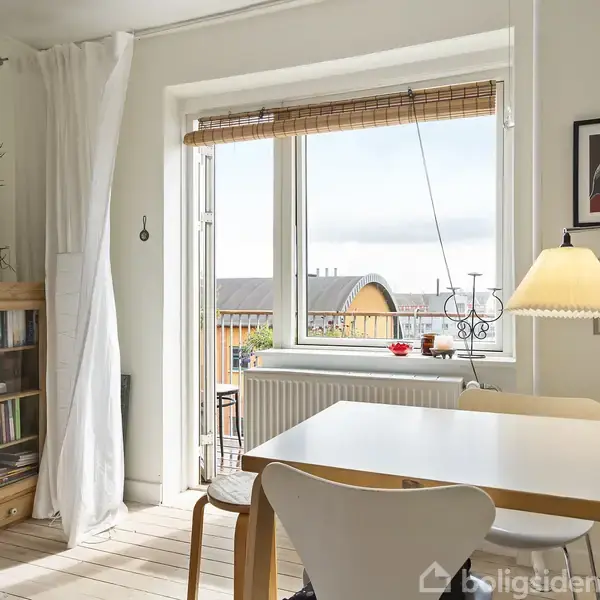 En lys spisestue med et bord og stole ved et vindue, hvor gardiner hænger til siden. En bogreol og planter står ved væggen. Udsigt til tagene udenfor.