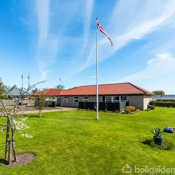 Et enfamiliehus med rødt tag står i en velplejet have. Et flag vajer på en høj flagstang ved siden af huset. Himlen er klar og blå.