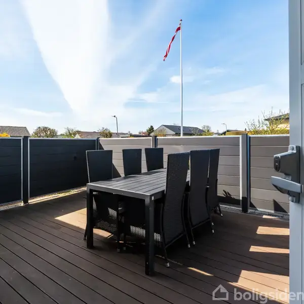 Terrassemøbler står på en træterrasse. Bordet er omgivet af seks højryggede stole. I baggrunden ses et hegn og en flagstang med et dansk flag i en rolig villakvarter.