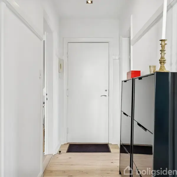 Dør: Hvid dør står i enden af en lys gang. Skoskab: Sort skoskab med stearinlys placeret ovenpå. Kontekst: Lyse vægge og trægulve skaber en minimalistisk indgang.