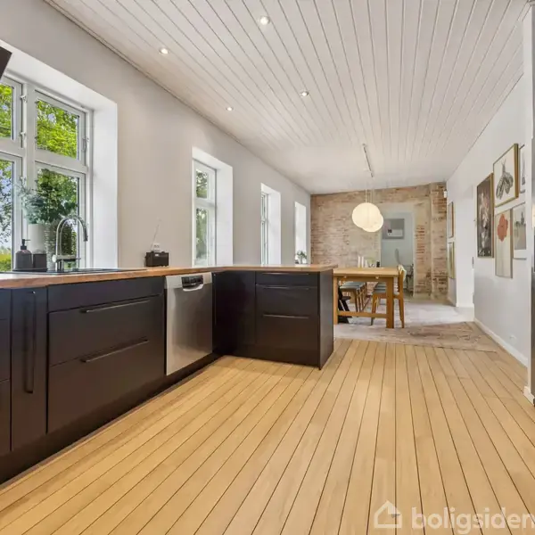 Køkken med sorte skabe står ved store vinduer. Et spisebord ses i baggrunden. Hvide lofter og lys træparket præger rummet, som er åbent og lyst.