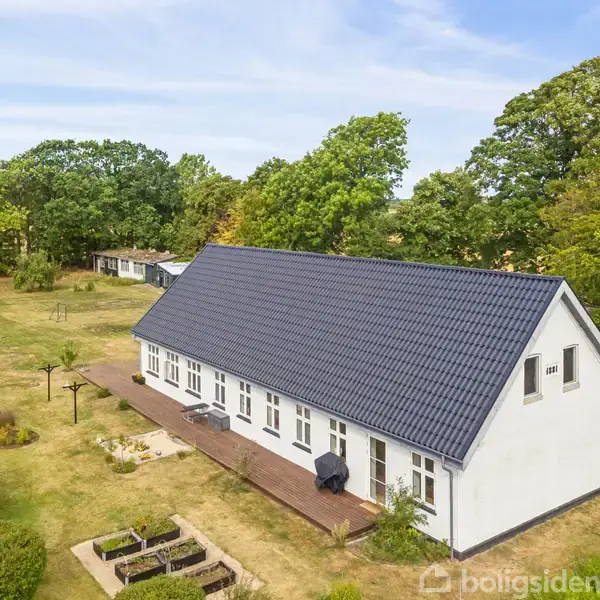En hvid bygning med mørkt tag står stille, omgivet af en stor have med træer og græsplæne. En legeplads og små blomsterbede vises i forgrunden.
