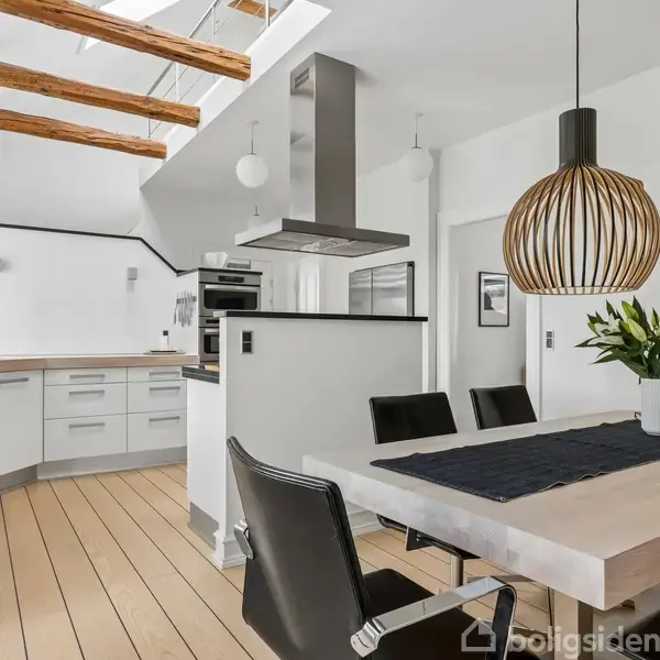 Et moderne køkken har en spisebord med sorte stole og en trælampe hængende over det. Køkkenet er lyst og rummeligt med hvide skabe og synlige loftsbjælker.