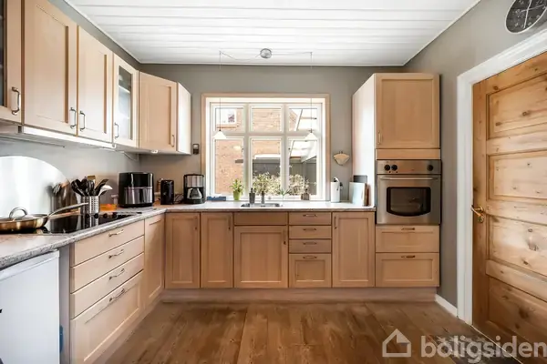 Køkken med lyse træskabe og grå bordplade; vindue over vask, ovn indbygget i højre skab. Trægulv, grå vægge, kaffe- og køkkenredskaber til venstre for vasken.