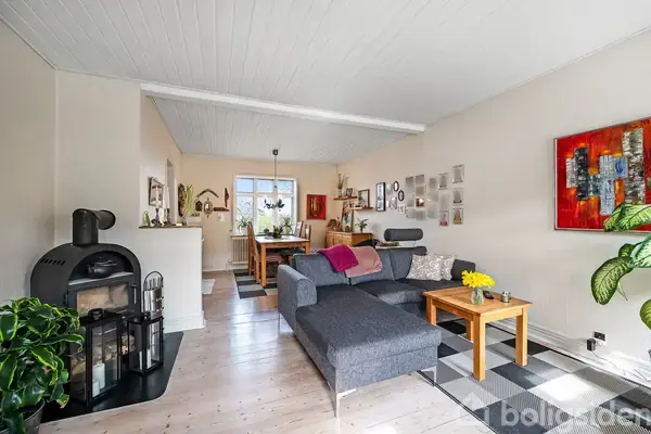 En stue med en grå sofa og et sofabord, placeret i et lyst rum med trægulv. I baggrunden er der et spisebord ved et vindue. Vægdekorationer og planter skaber en hyggelig atmosfære.