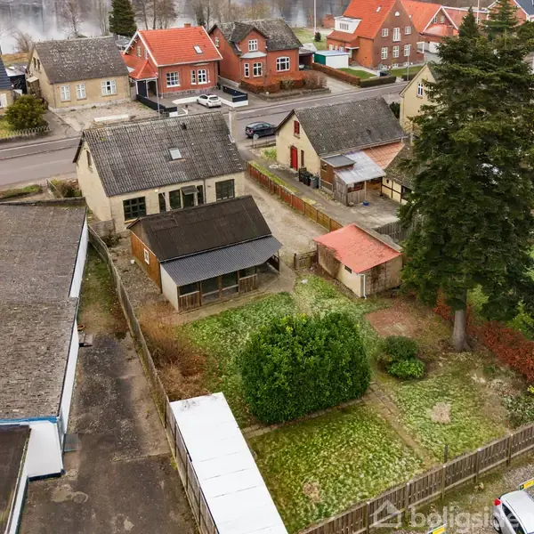 Et luftfoto viser en række huse med grå og røde tage, omgivet af en gade og parkerede biler. Haven i forgrunden har en stor busk og træer.