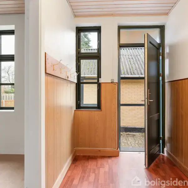 En åben dør leder ud til en flisebelagt gårdsplads. Indenfor er der trægulv og træpaneler på væggene. Til venstre fører en åbning til et andet rum med vinduer.