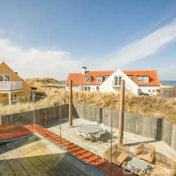 Terrasse med kurvestole og rundt bord omgivet af hegn, placeret i nærheden af gule og hvide huse med røde tage. Sandklitter og hav i baggrunden under en lys blå himmel.