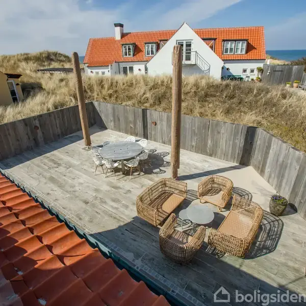 En træterrasse med et bord og fire stole er omkranset af et hegn. Der er to høje træstolper. Baggrunden viser huse med røde tage og en overskyet himmel.