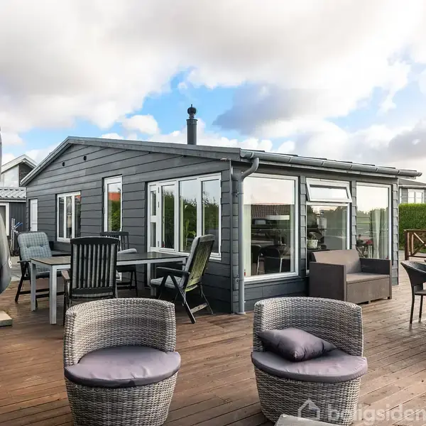 Gråt træhus med store vinduer står stille på en stor terrasse fyldt med havemøbler. Omgivet af grønne buske under en skyet blå himmel.