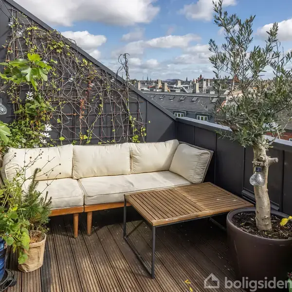 En hvid sofa står på en træterrasse med en sort metalramme, omringet af planter i krukker. Bagved er et gitter med klatreplanter, og i baggrunden ses byens tage.