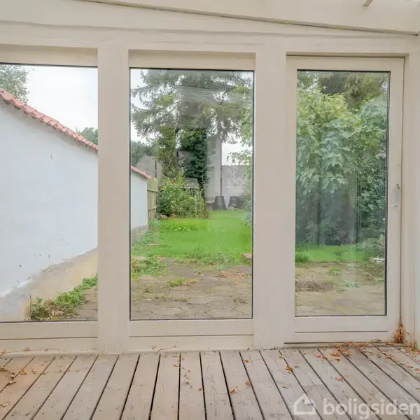 Glasparti med udsigt over en lille terrasse og frodig have. Hvidmalede vægge indendørs, trægulv med blade. Udendørs vises en græsplæne omgivet af grønne buske og træer.