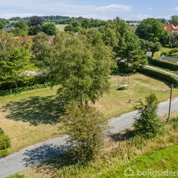 Græsplæne med enkelte træer står stille i et parcelhuskvarter omgivet af hække og veje. I baggrunden ses flere huse med haver og frodig beplantning.