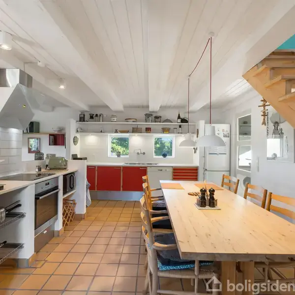 Spisebord med træstole er centreret i et lyst, moderne køkken-alrum, omgivet af røde køkkenskabe, hvide vægge, flisegulv og en trætrappe. Køkkenudstyr er synligt på bordet.