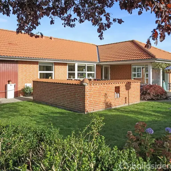 Et rødstenshus med rødt tegltag står stille i en velplejet have. En græsplæne omgiver huset, mens blomstrende buske strækker sig mod solen under en klar, blå himmel.
