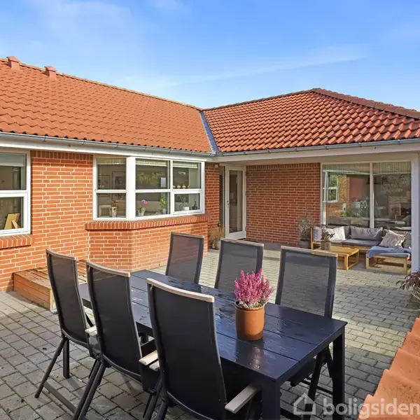 Hus med røde mursten og tegltag. Havebord med stole står på flisebelagt terrasse. I baggrunden er der en dør og et sofahjørne med puder og tæpper i en solrig have.