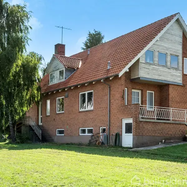 En rød murstensvilla med skråt tag står på en grøn græsplæne omgivet af træer. Huset har flere vinduer og en udvendig trappe, der fører til en dør.