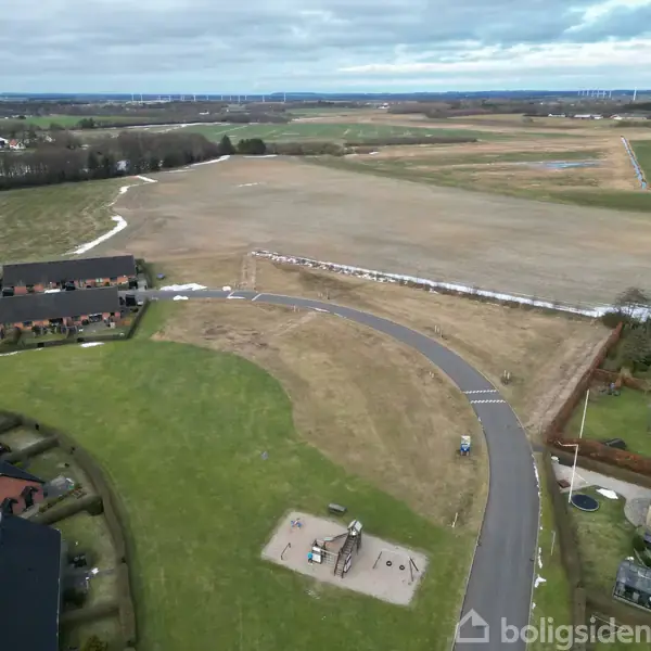 Legeplads placerer sig på en græsplæne ved siden af en vej med landområde i baggrunden. Den er omgivet af huse og åbne marker under en overskyet himmel.