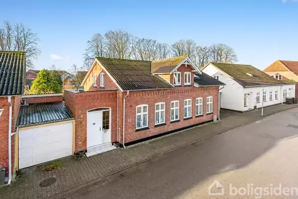 Et rødt murstenshus står stille i en rolig gade, flankeret af et garageanlæg til venstre og et hvidt hus til højre. Træer ses i baggrunden.