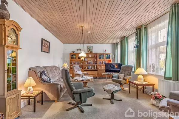 En stue med sofaer og lænestole står omkring et sofabord. I baggrunden er en reol fyldt med nips, og et TV står til højre. Vinduer med grønne gardiner slipper lys ind.