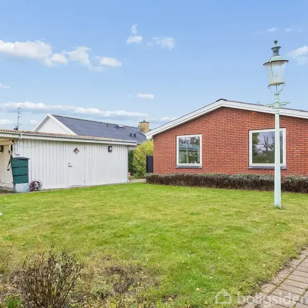 En rød murstensvilla med vinduer, står stille omgivet af en grøn have. En flisebelagt sti løber langs huset, og en hvid lygtepæl står i plænen.