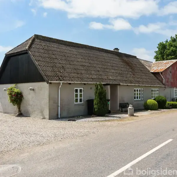 En traditionel, firlænget gård med gråt murværk og teglstenstag. Stille beliggende ved en landevej, omgivet af grusbelagt gårdsplads og grønne buske. Klar og solrig himmel.